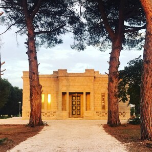A building with a central entrance, flanked by two large trees, and a gravel path leading to it. - Interior Designed "Mini Castle" with Pool in Puglia (Francavilla Fontana)