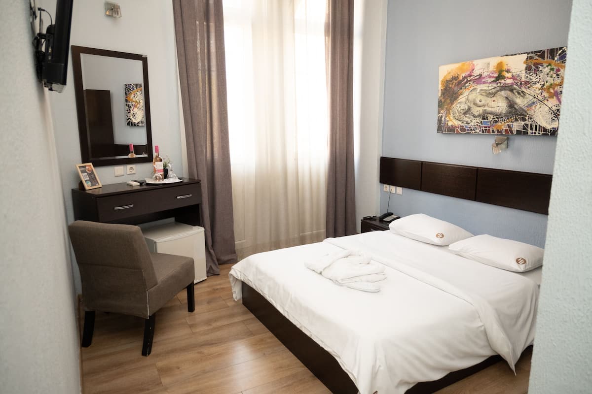 A hotel room with a bed, a desk, a chair, a dresser, a painting on the wall, and a window with curtains.