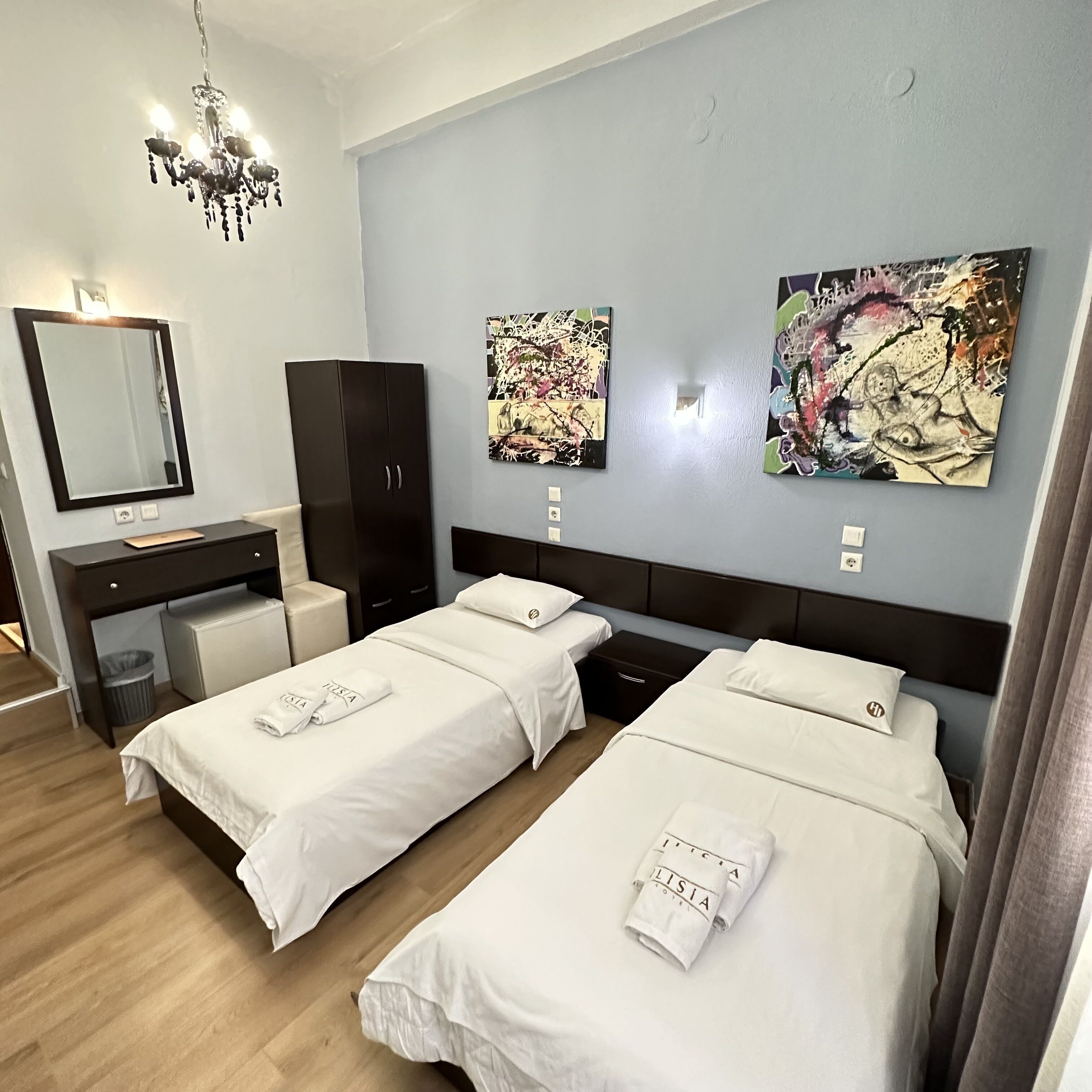 A hotel room with two beds, a desk, a wardrobe, and two abstract paintings on the wall.