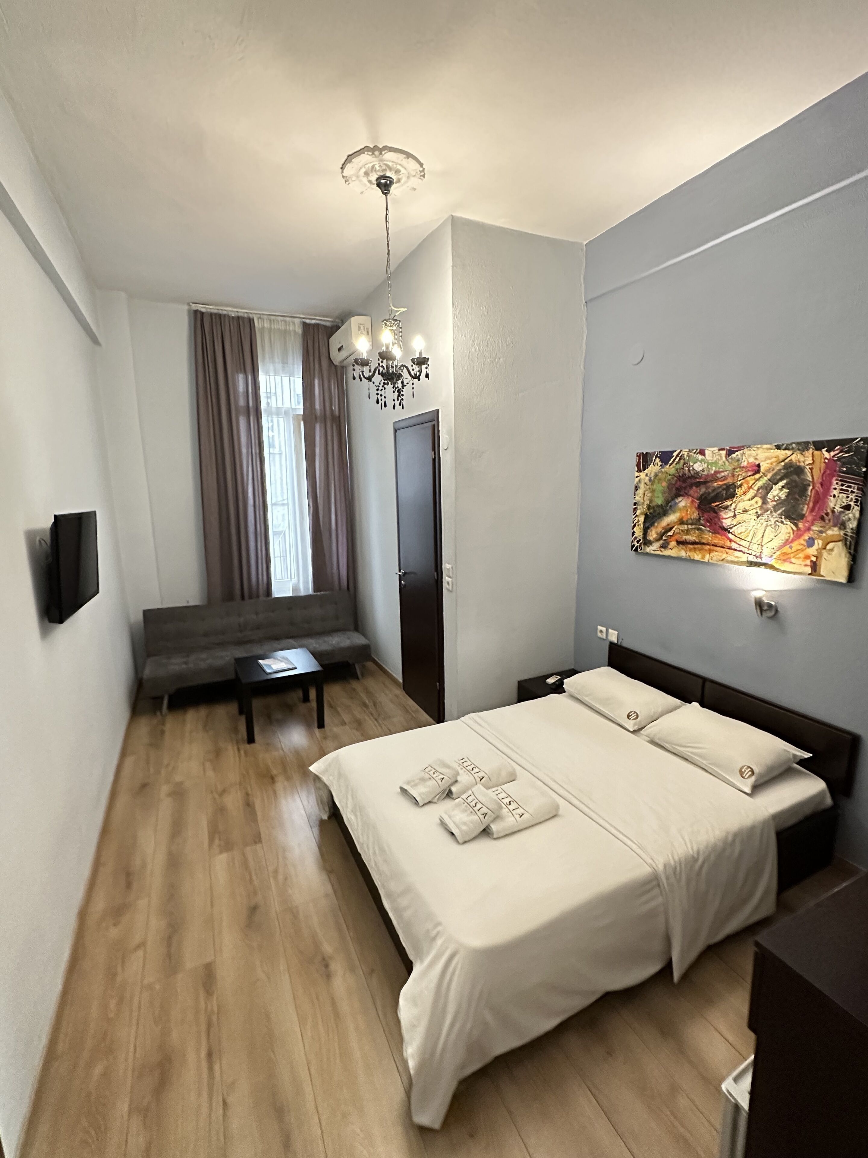 A modern hotel room with a bed, a sofa, a small table, a chandelier, and a painting on the wall.