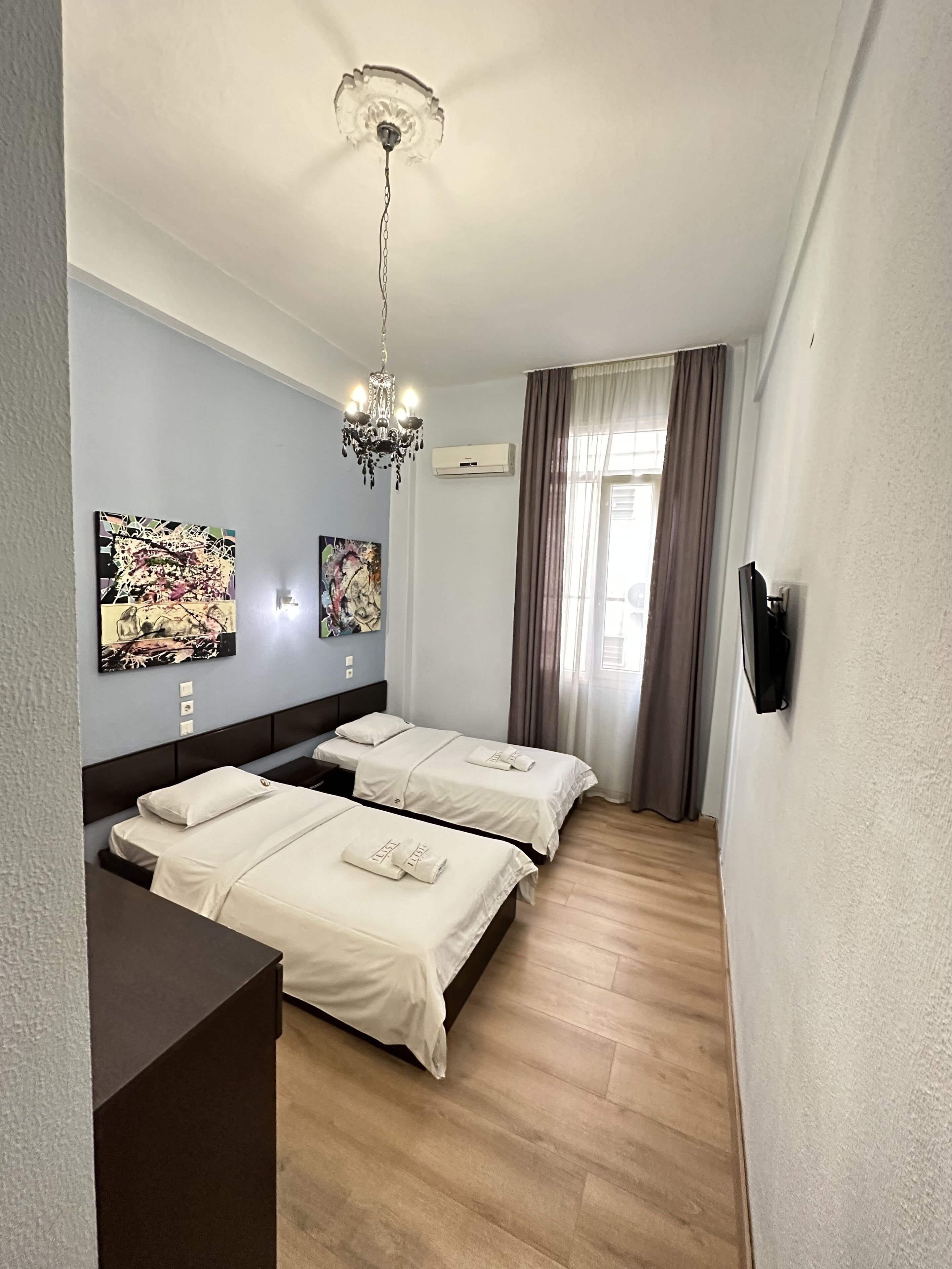 A hotel room with two beds, a chandelier, artwork on the wall, and a window with curtains.