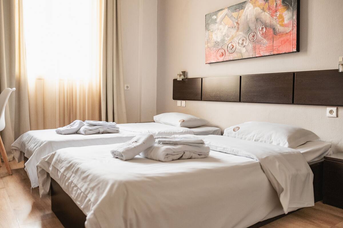 A hotel room with two beds, a painting on the wall, and a window with curtains.
