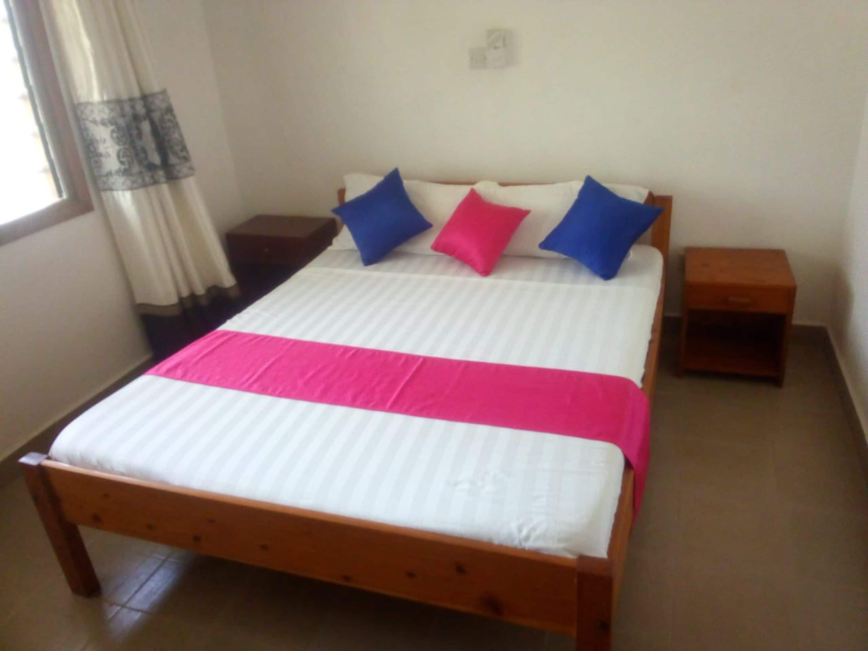A bed with a white mattress, a pink and white striped blanket, and blue and pink pillows. There is a wooden nightstand to the right of the bed.