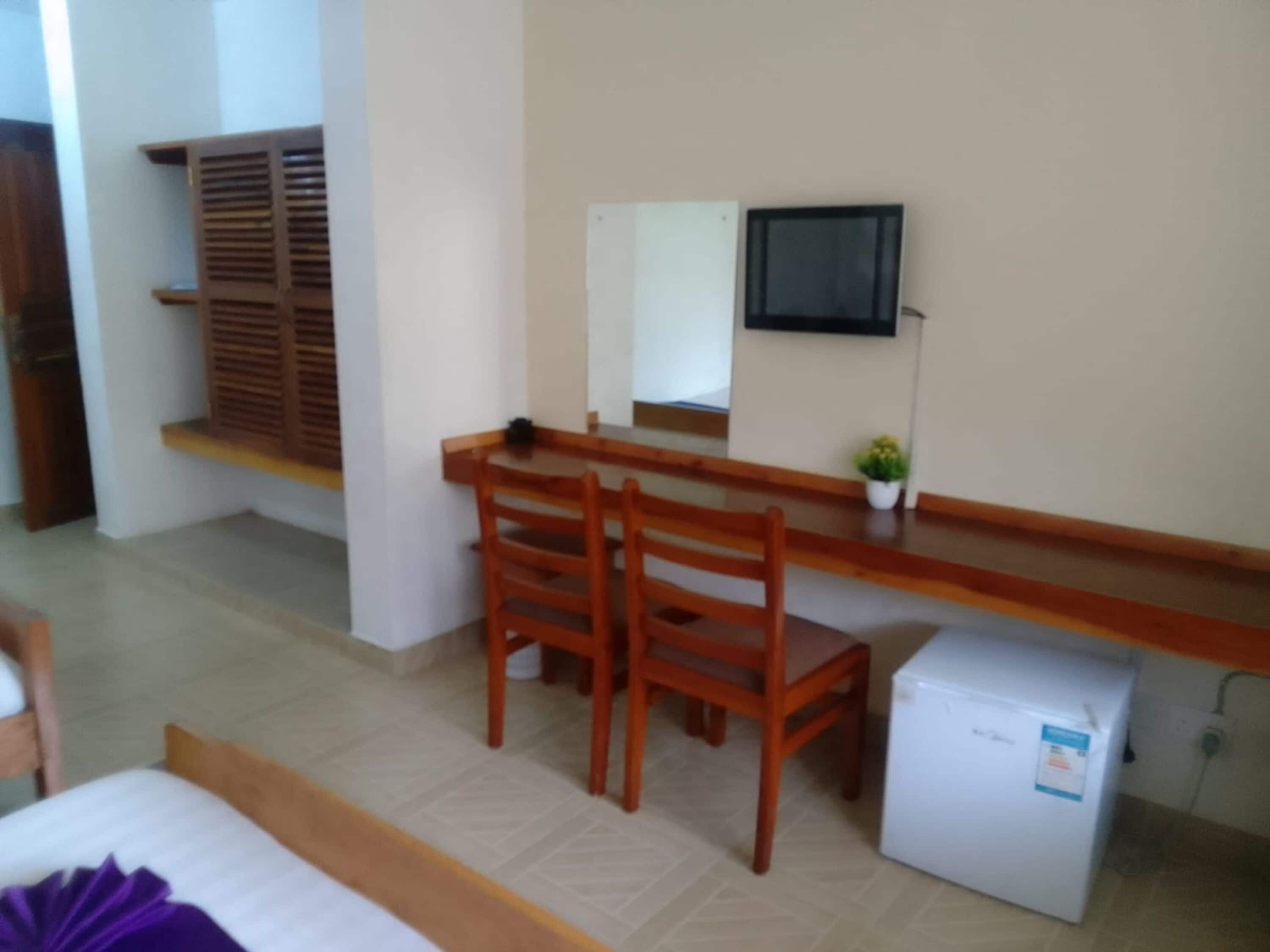 Ein kleines Zimmer mit einem Holztisch, zwei Stühlen, einer kleinen Kühlbox und einer Topfpflanze.