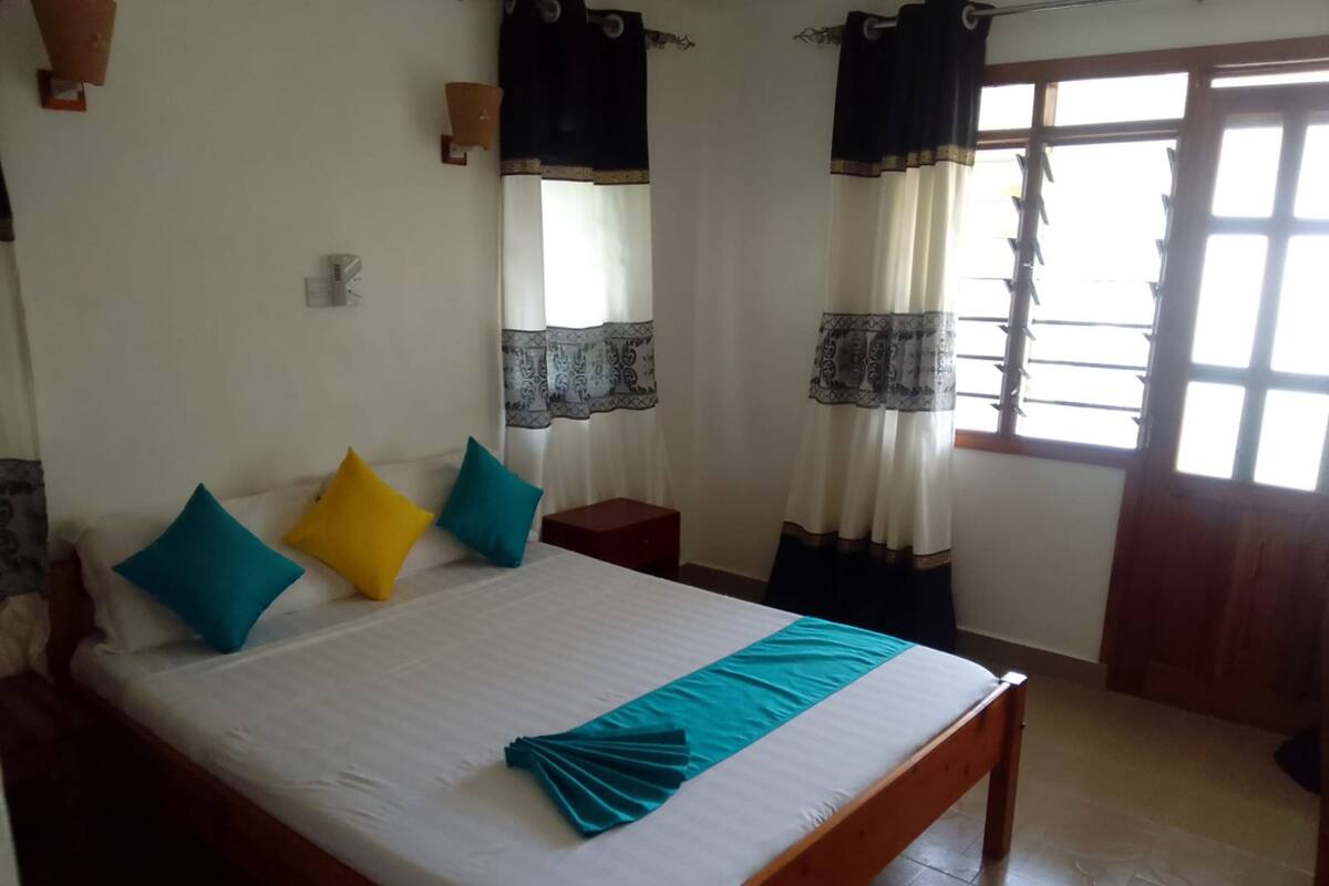 Un dormitorio con cama de madera, ropa de cama blanca y almohadas coloridas. Hay dos ventanas con cortinas, una de ellas con un diseño decorativo.