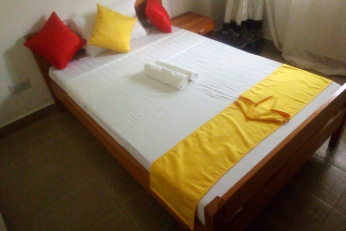 Una cama bien hecha con sábana blanca, manta amarilla y dos almohadas, una roja y otra amarilla.