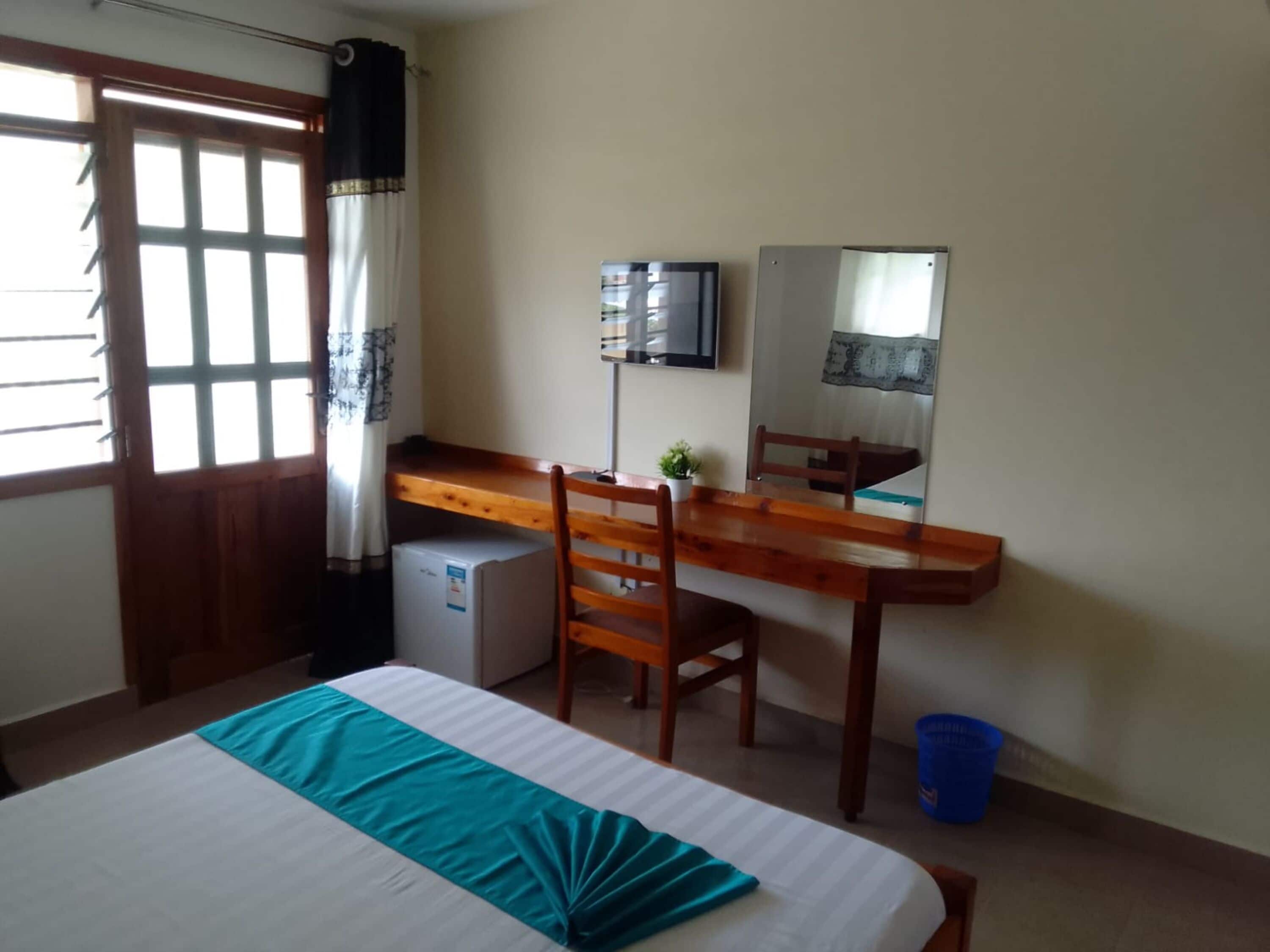 A room with a bed, a desk, a chair, a small refrigerator, a television, and a window with curtains.