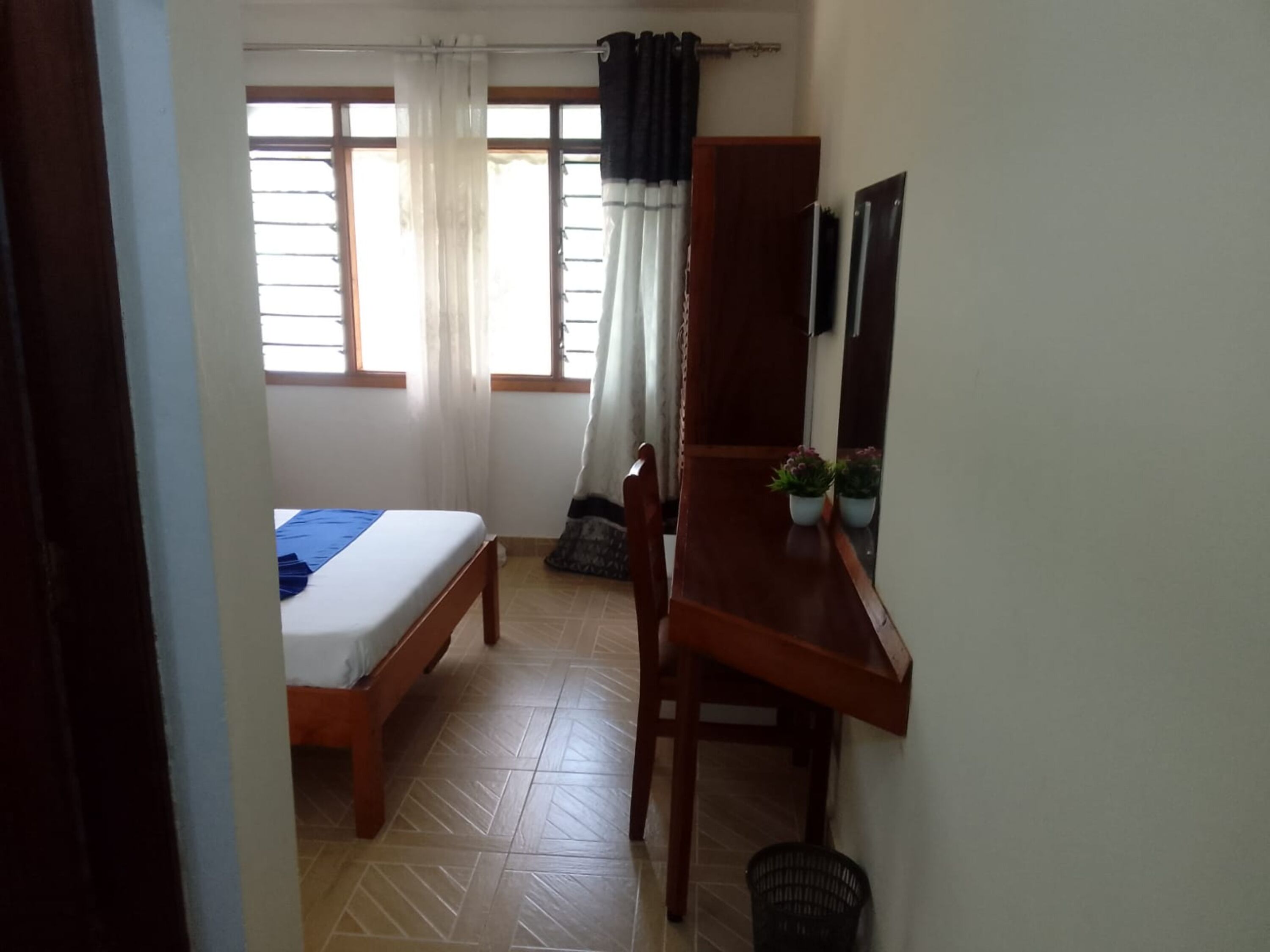 A room with a bed, a desk, and a chair. There are windows with curtains, a potted plant, and a mirror on the wall.