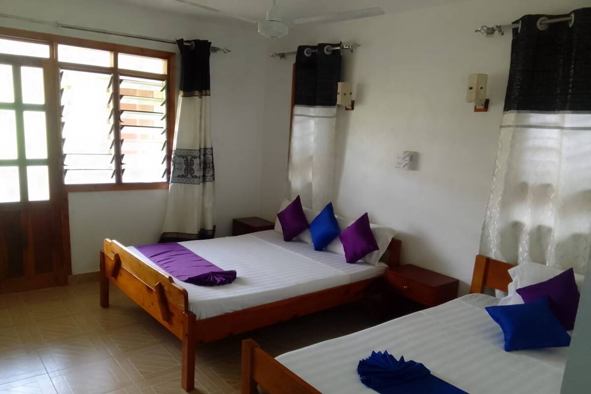 Una habitación con dos camas de madera, una ventana con cortinas, una mesita de noche y una lámpara fijada a la pared.