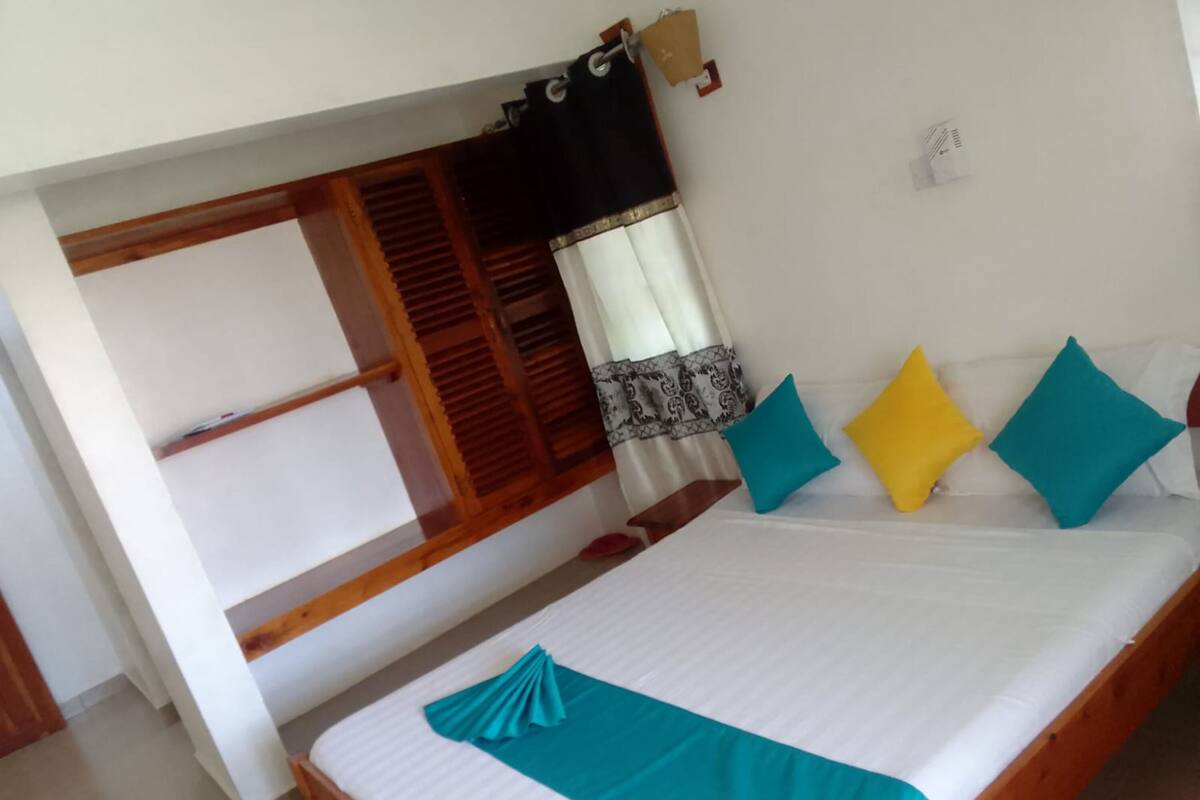 Una cama individual con edredón y almohadas color turquesa, cabecero de madera, y una ventana con contraventanas y cortinas de madera.