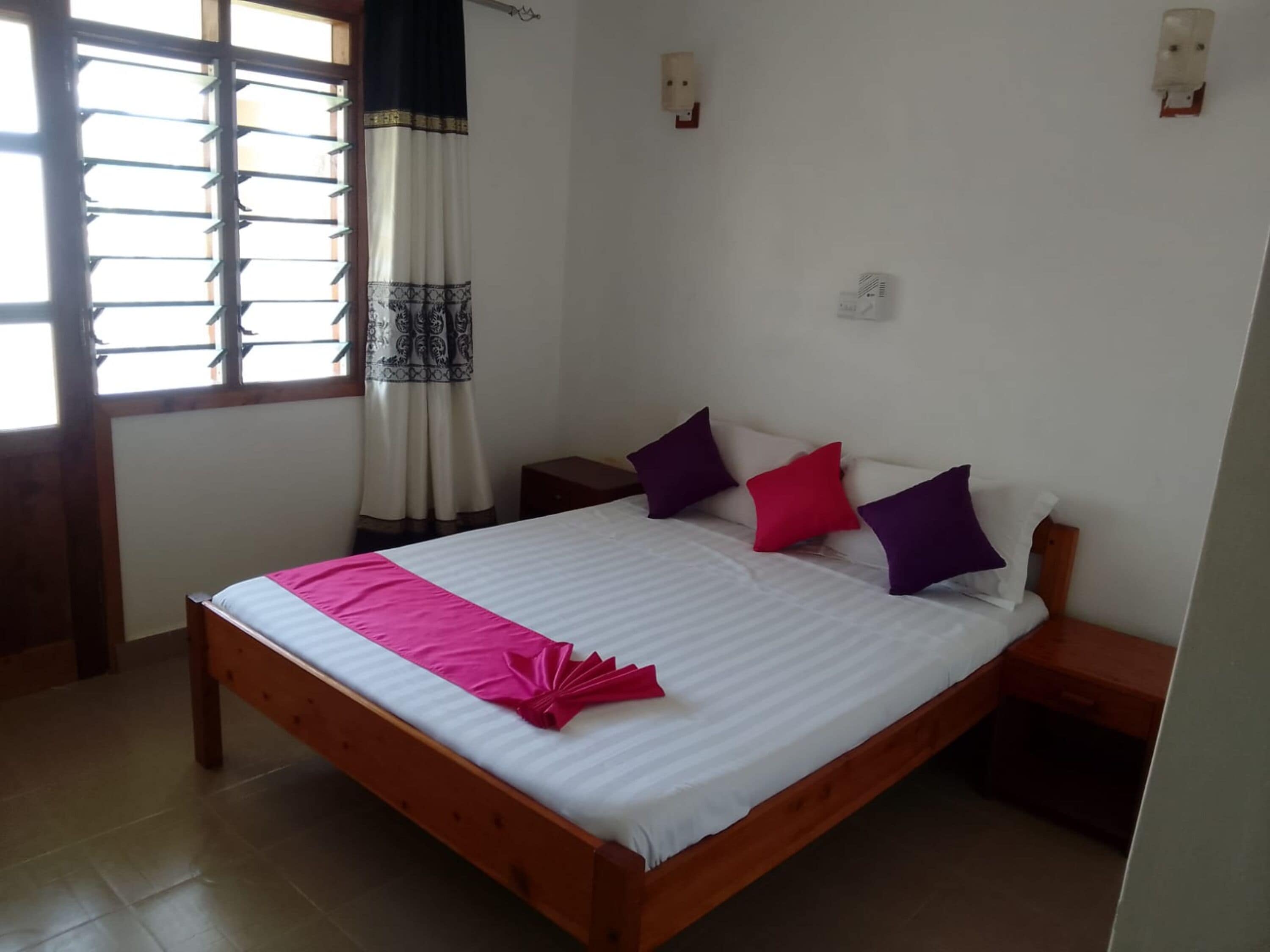A neatly made bed with a pink and white bedspread, a wooden headboard, and multiple pillows in purple and red.