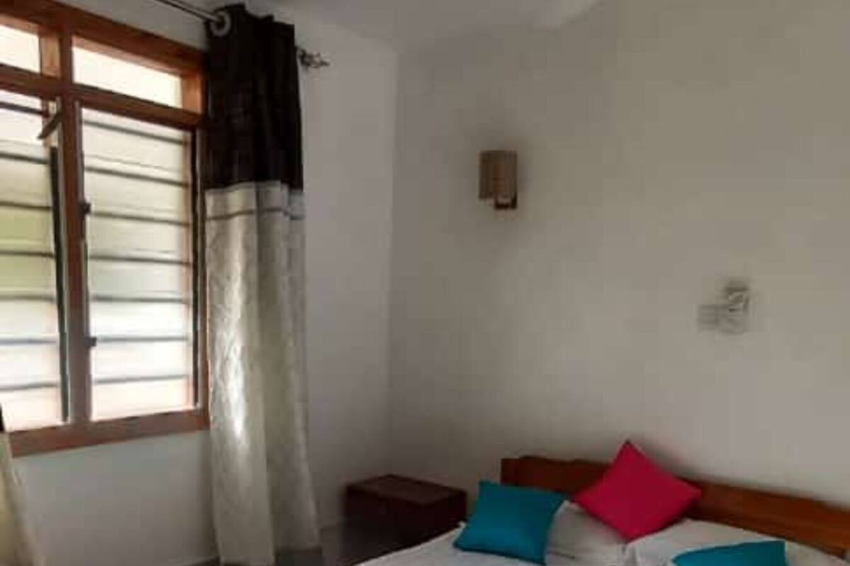 Un dormitorio con cama, ventilador de techo, ventana con cortinas y una mesita de noche.