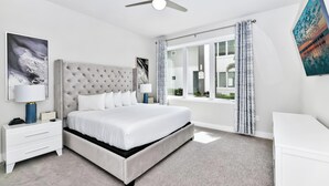 Ein modernes Schlafzimmer mit einem bettenhohen, gesteppten Kopfteil, weißen Nachttischen, einem Deckenventilator und großen Fenstern mit Vorhängen.