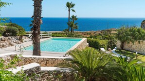 A rectangular pool with a stone wall, surrounded by palm trees and greenery, overlooking a clear blue sea. - Villa with pool, sea front location , Plemmirio, DiCasaInSIcilia (Siracusa)