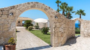 A stone archway leading to a building with a tiled roof, surrounded by palm trees and a clear blue sky. - Villa with pool, sea front location , Plemmirio, DiCasaInSIcilia (Siracusa)