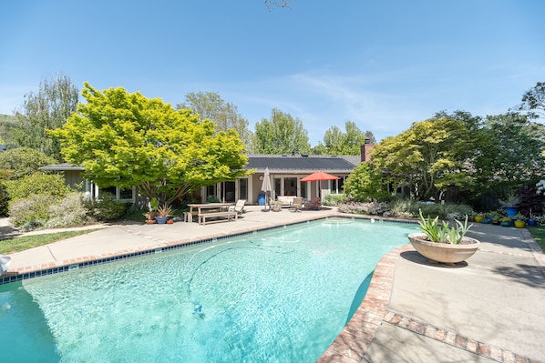 Una piscina con bordeado de baldosas, rodeada por un patio con mobiliario exterior y plantas en macetas.