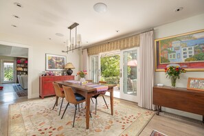 Un comedor con una mesa de madera, sillas y un tapete estampado. Se observa una zona de cocina con un armario rojo y un cuadro en la pared.