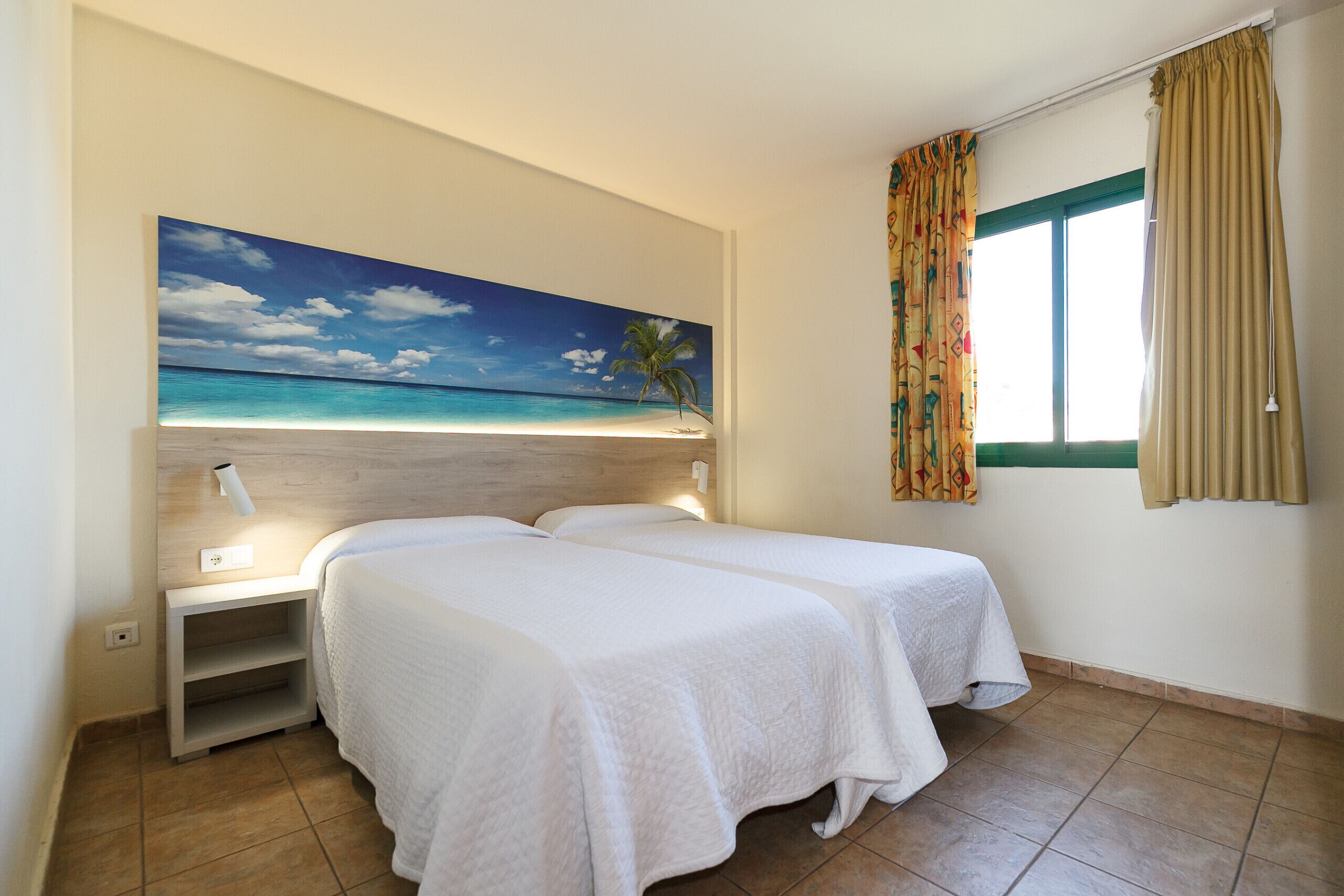 Une chambre d’hôtel avec deux lits, un tableau représentant une scène de plage, une fenêtre avec des rideaux et une table de chevet avec une lampe.