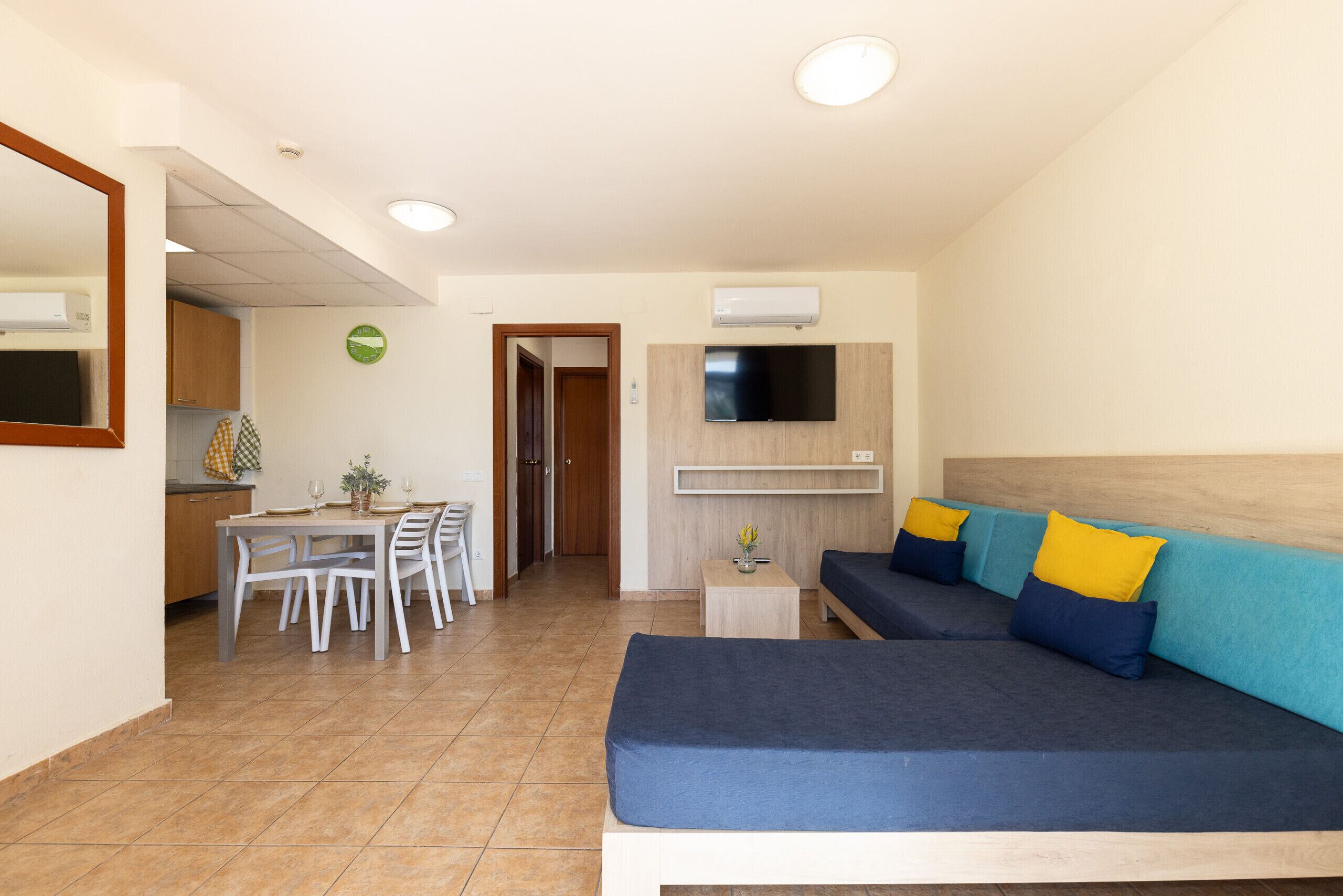 Una sala de estar moderna con un sofá, una mesa de comedor con sillas, un televisor montado en la pared y una pequeña mesita de noche con una planta.