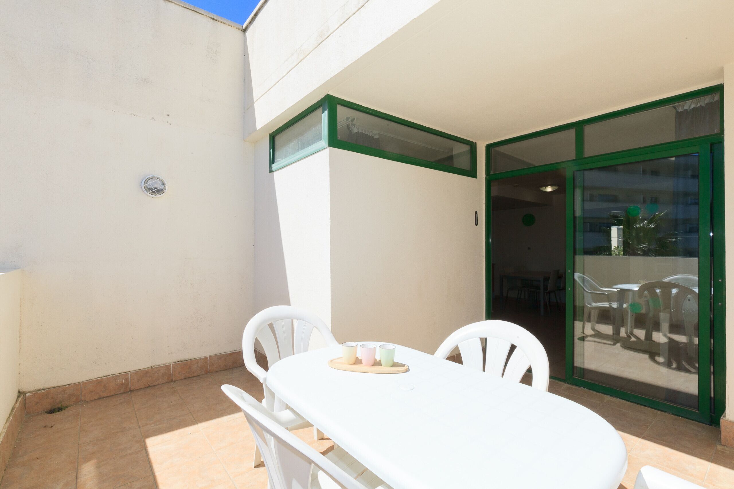 Una mesa blanca con sillas y una mesita con tazas, frente a una pared blanca con una puerta de marco verde.