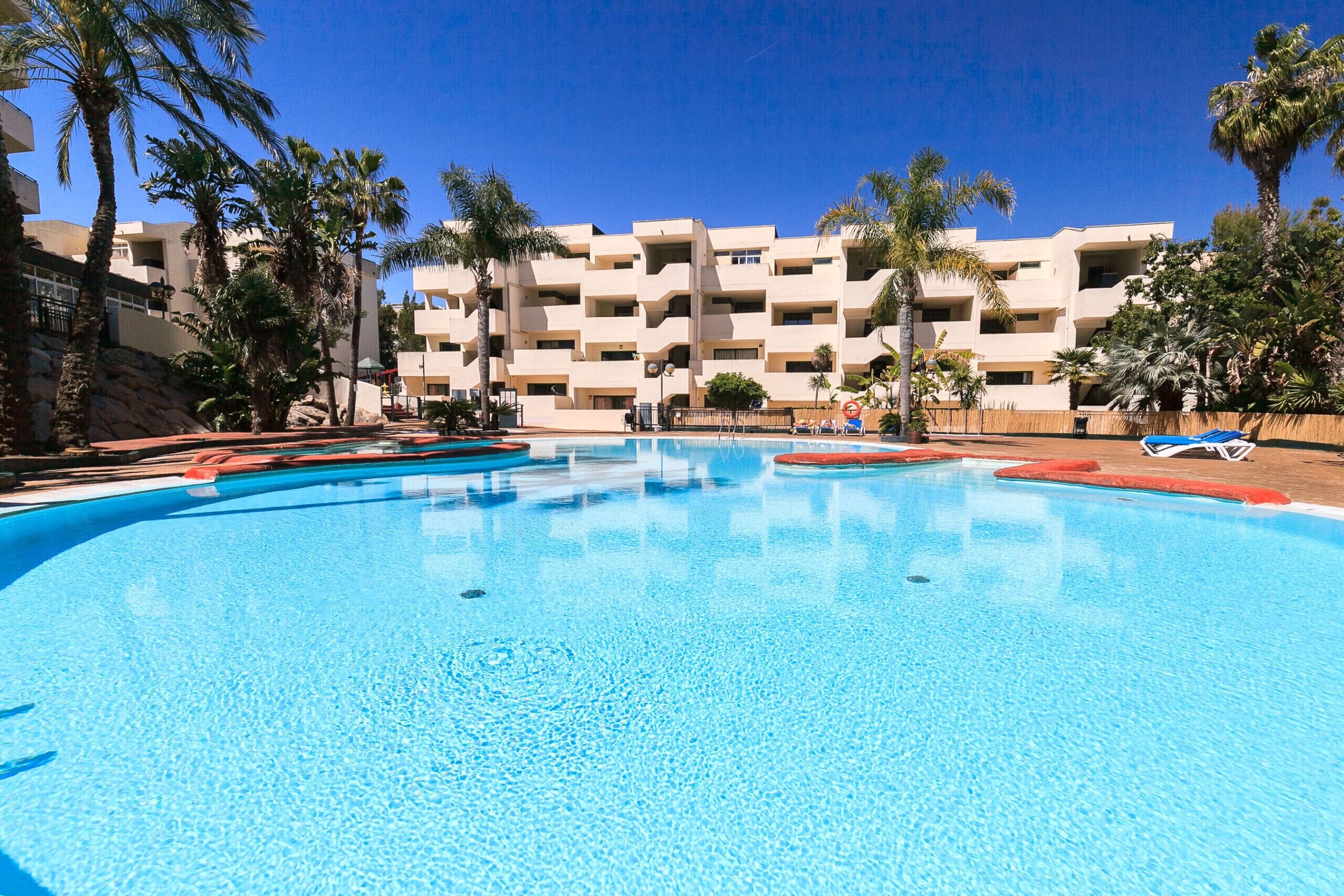 Ein Swimmingpool mit klarem, blauem Wasser, umgeben von Palmen und einem mehrstöckigen Gebäude mit Balkonen.