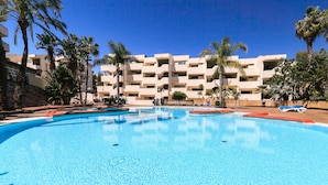 A swimming pool with clear blue water, surrounded by palm trees and a multi-story building with balconies. - FESTIVAL VILLAGE APARTMENTS (Salou)