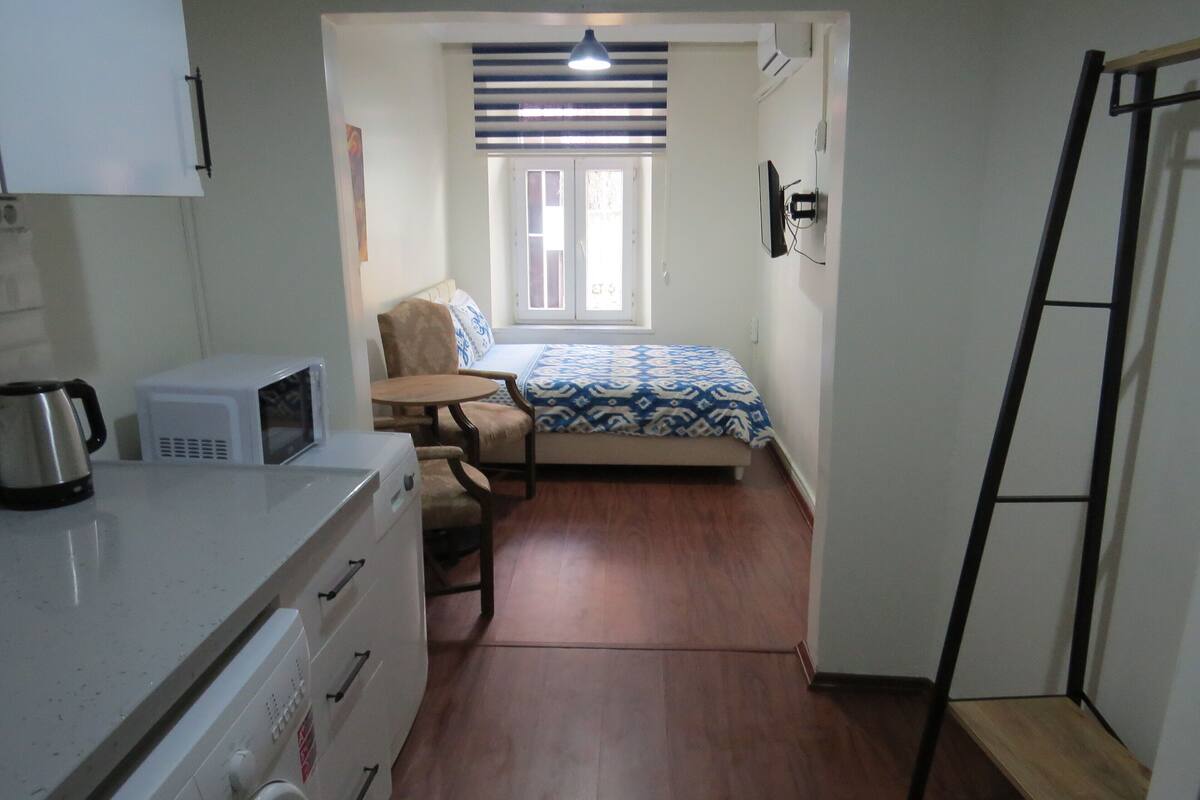 A compact room with a kitchenette, a bed with patterned bedding, a window with blinds, and a wooden floor.