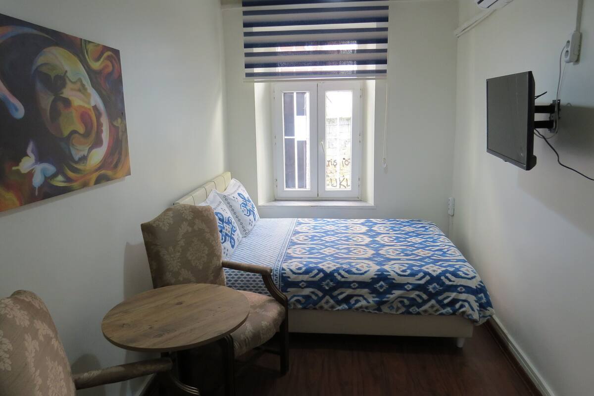 A small, well-lit room with a bed, a chair, a table, and a window with blinds.