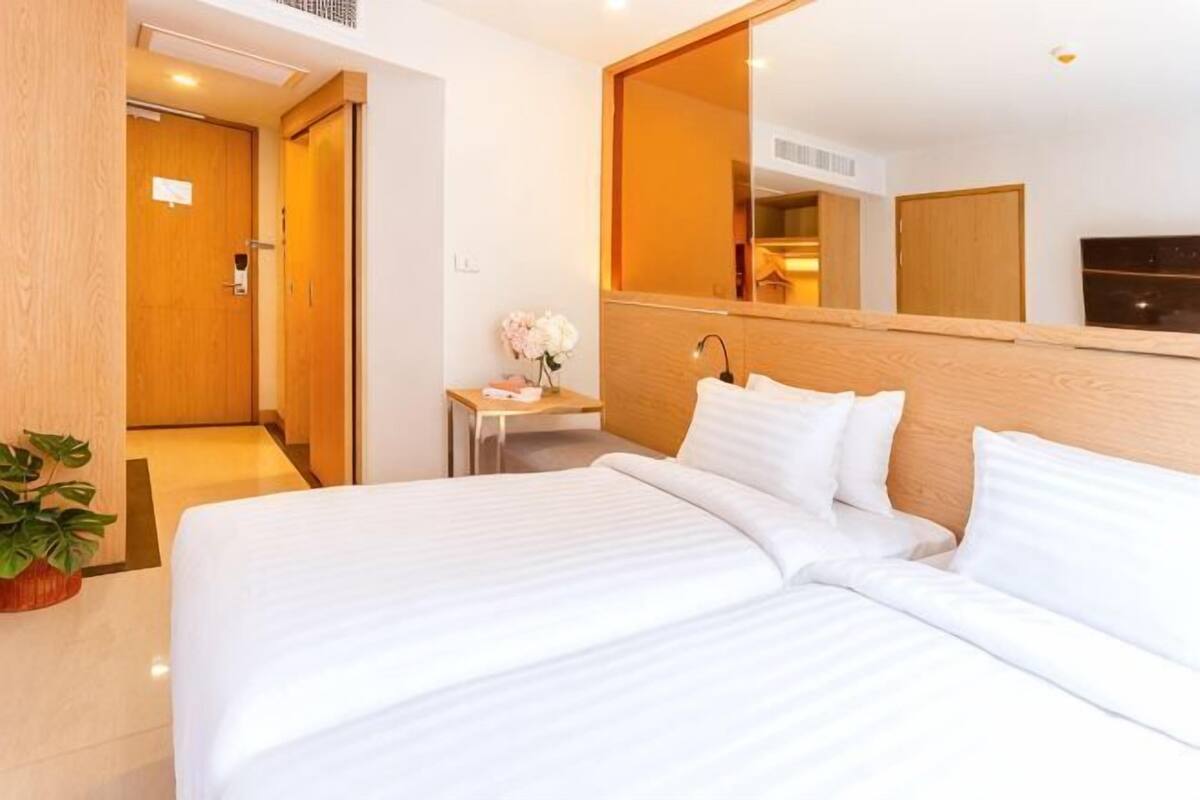 Una habitación de hotel con una cama grande, una mesita de noche con un jarrón, un espejo sobre la cama y una puerta de madera.