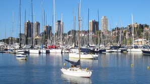 A marina with multiple sailboats docked, a city skyline in the background, and calm water reflecting the boats.