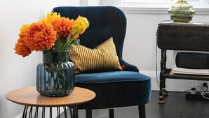 A modern living room with a dark blue armchair, a small round table with a vase of orange flowers, a yellow striped pillow, and a lamp on a side table.