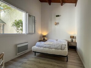Un dormitorio con cama, dos lámparas de noche, un radiador, una ventana con vistas a la vegetación y una planta pequeña en la pared.