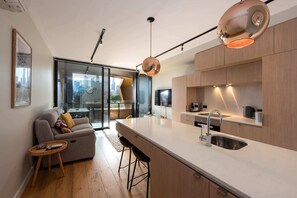 A modern kitchen with a large island, stainless steel sink, and wooden cabinets. There is a sofa with a small side table and a city view through large windows. - 2-bed in South Melbourne w/ Beautiful City Views (South Melbourne)