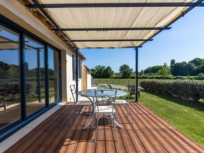 Una terraza de madera con una mesa y sillas, ventanas amplias y vistas a un jardín.