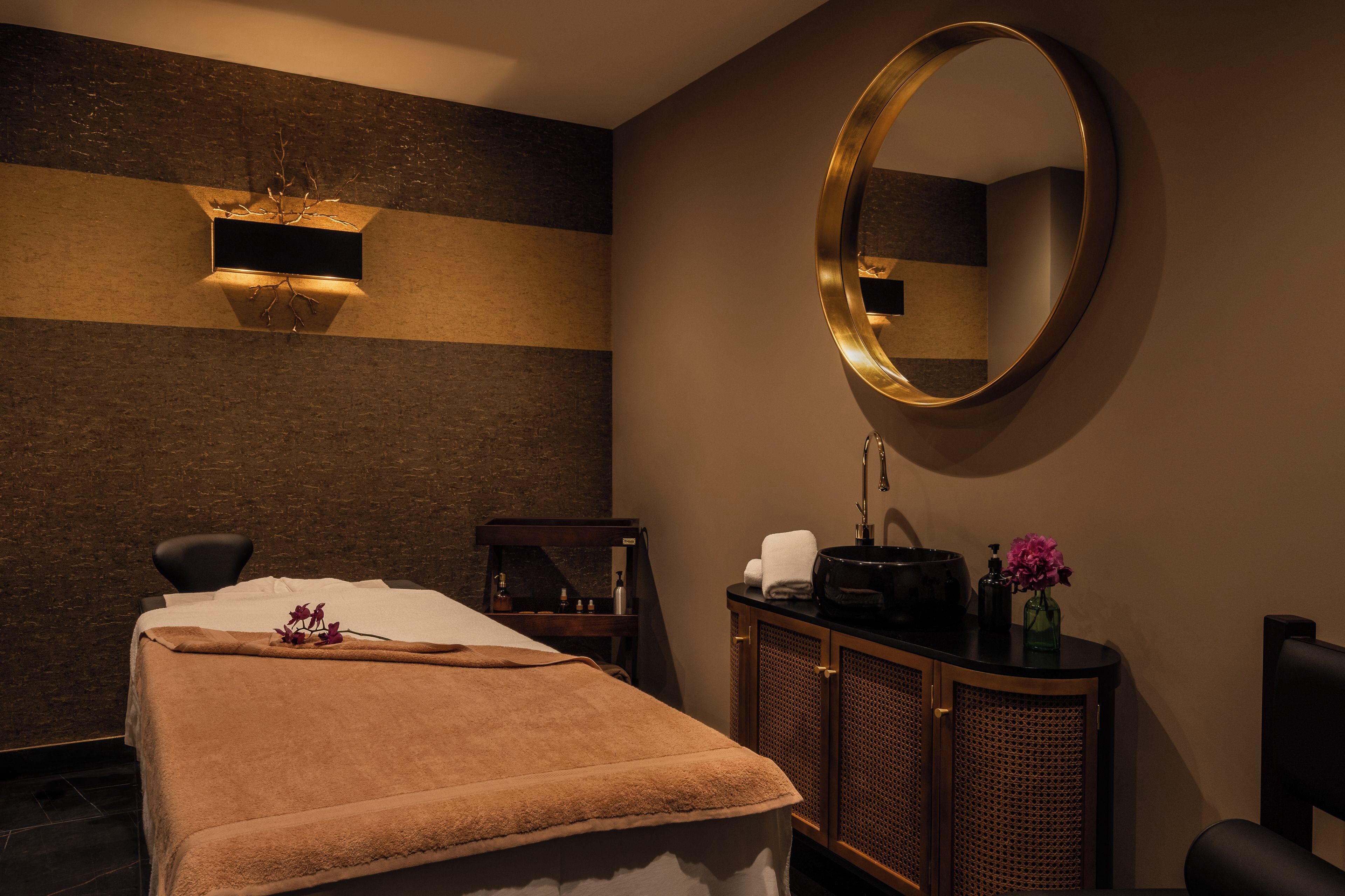 A spa room with a massage table, a large mirror, a cabinet with toiletries, and a wall-mounted light fixture.