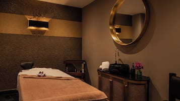 A spa room with a massage table, a large mirror, a cabinet with toiletries, and a wall-mounted light fixture.