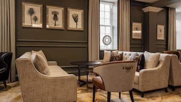 A living room with a beige sofa set, a round coffee table, and a patterned rug. There are framed botanical prints on the wall and a window with curtains.