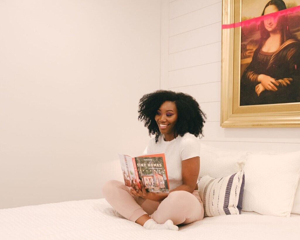Iemand zit op een bed te lezen, in een kamer met een witte muur en een schilderij boven het bed.