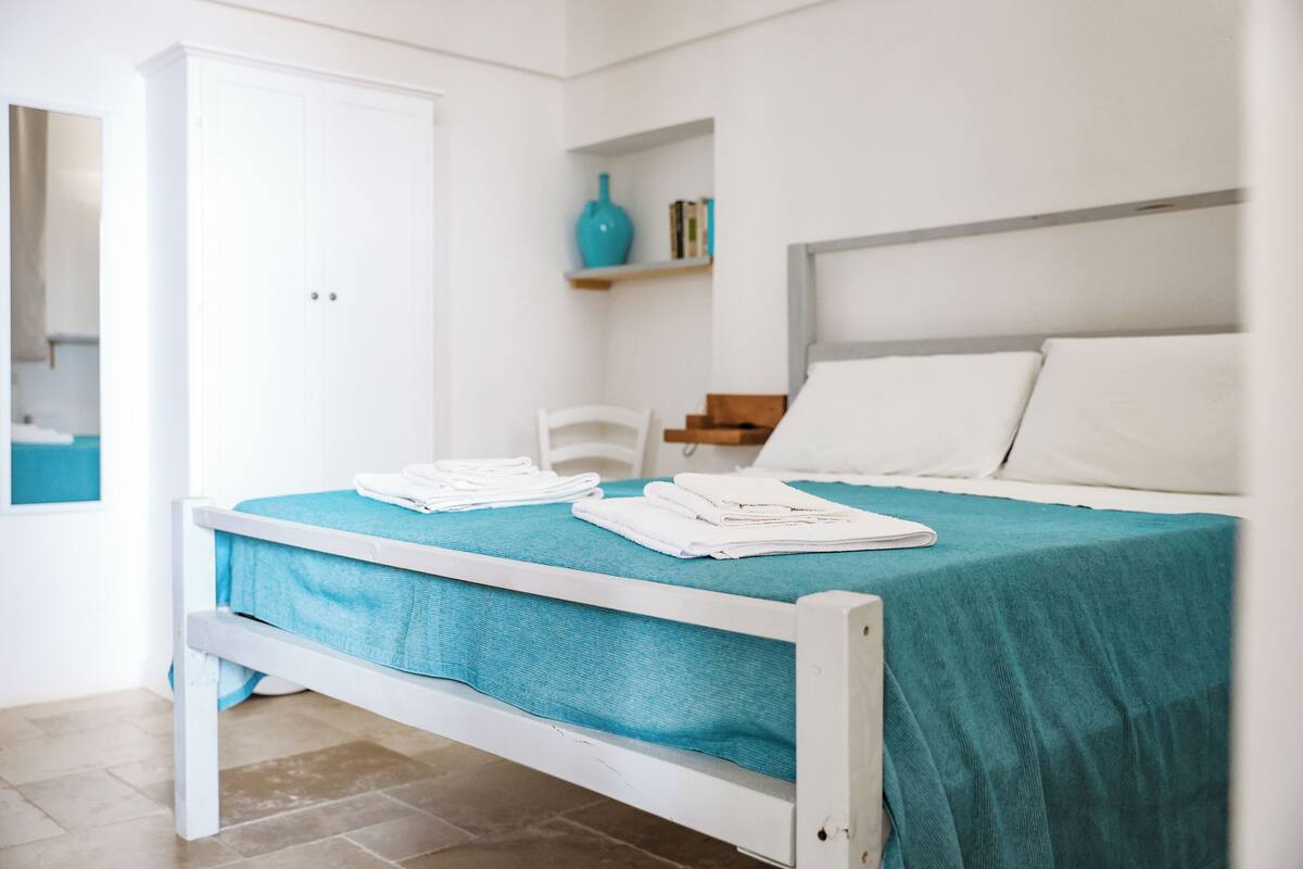 Un lit bien fait, recouvert d’une couvre-lit turquoise et agrémenté d’oreillers blancs, une armoire blanche et une petite étagère avec des livres et un vase bleu.