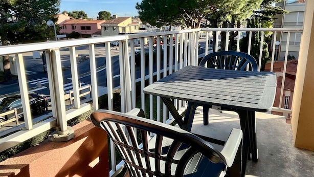 A balcony with a table and chairs, overlooking a street with parked cars and buildings. - Cozy Studio for 4 with A/C, Pools & Parking - 400m from Beach, 800m to Town Centre (Argelès-sur-Mer)