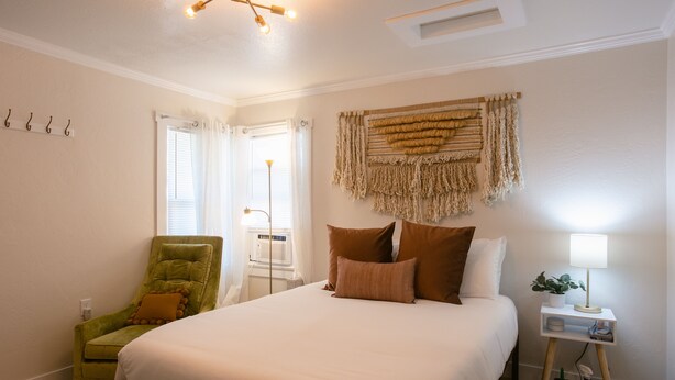 A bedroom with a large bed, a green armchair, a bedside table with a lamp, and a wall-mounted tapestry. - Enjoy the tranquility of a scenic riverside neighborhood in Gold Hill. This area is peaceful, offers beautiful views, and is a convenient stopover location. (Gold Hill)