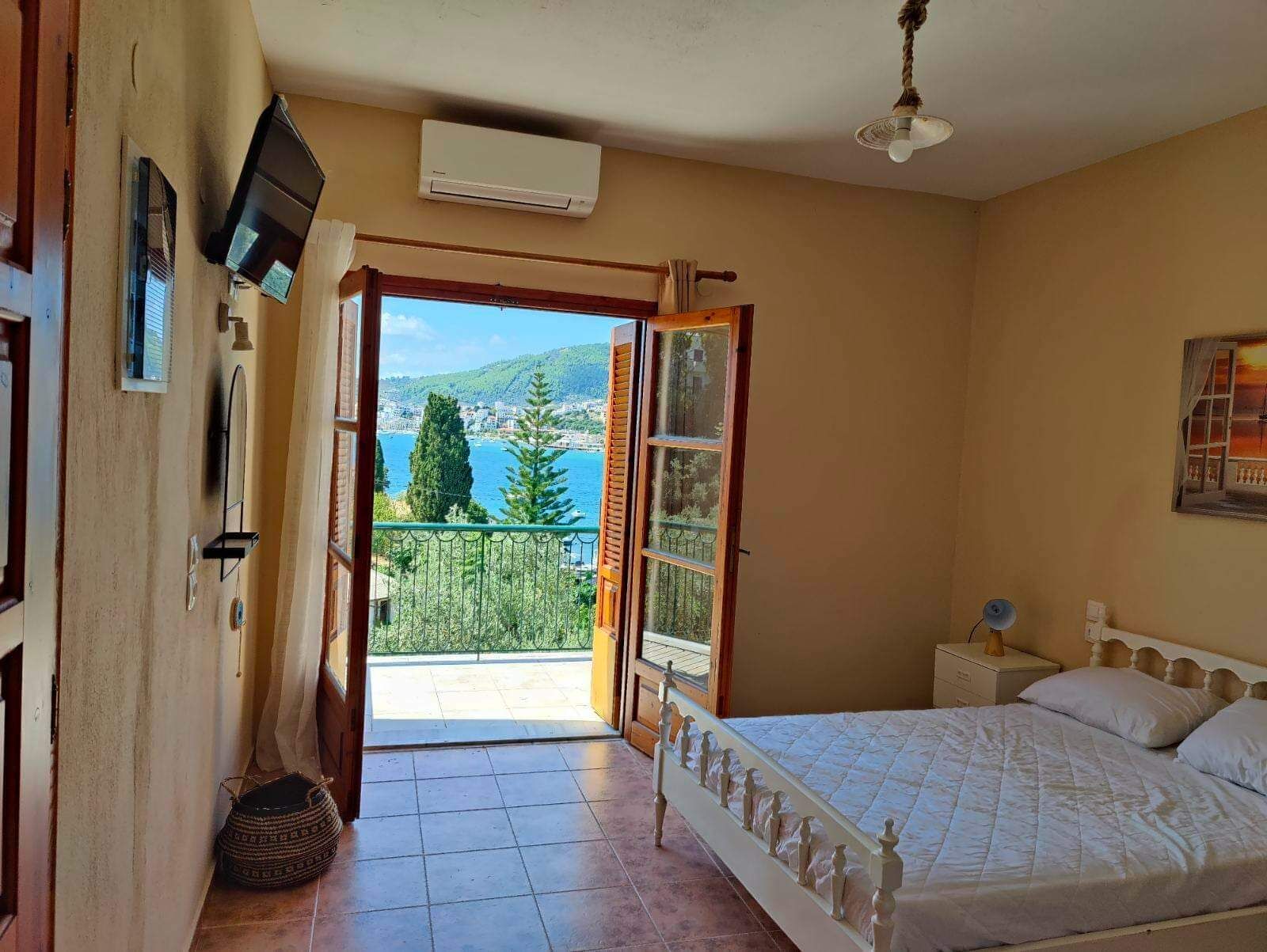 A bedroom with a bed, a nightstand, a lamp, a TV, and a balcony with a view of the sea and hills.