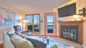 Una sala de estar con sofá, sillón, chimenea, televisor y ventanas amplias.