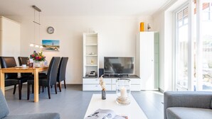 A modern living room with a dining area, a flat-screen TV, and a large window. - Großzügig. Garten. Zentral. ID: Mbs/22 (Westerland)