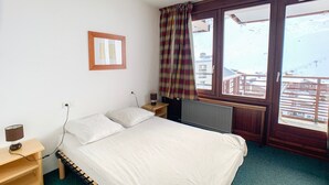 A bedroom with a bed, a nightstand, a lamp, a window with a view of buildings, and a curtain. - Comfortable apartment near slopes and shops with balcony, pets allowed (Tignes)