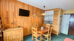 A compact room with a wooden wall, a dining table with four chairs, a small cabinet, a television mounted on the wall, and a door to the right. - Comfortable apartment near slopes and shops with balcony, pets allowed (Tignes)