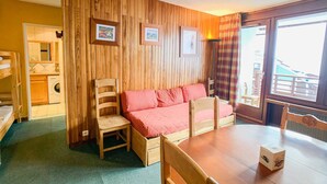 A cozy room with a wooden wall, a bed with red bedding, a wooden table, and a chair. There is a window with curtains and a view of buildings outside. - Comfortable apartment near slopes and shops with balcony, pets allowed (Tignes)