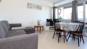 A modern living room with a grey sofa, a dining table with four chairs, a small side table with a vase, and a large window with curtains. - Charming 1-Bedroom Apartment, Balcony, WiFi, Near Thermal Baths, 2-Star Rating (Balaruc-les-Bains)