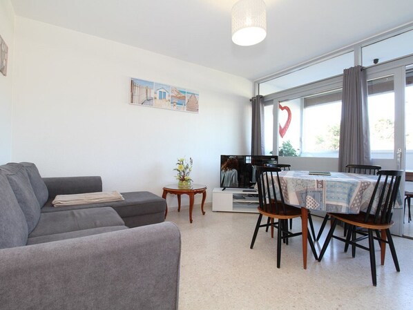 Un salon moderne avec un canapé gris, une table à manger avec quatre chaises, une petite table d’appoint avec un vase et une grande fenêtre avec des rideaux.