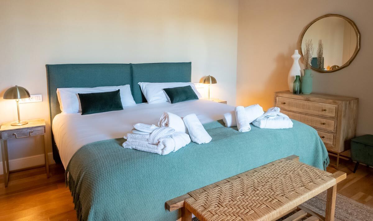 Un lit bien fait, recouvert d’une couvre-lit turquoise et agrémenté d’oreillers blancs, une table de chevet en bois avec une lampe, un banc en osier et une commode avec un miroir.